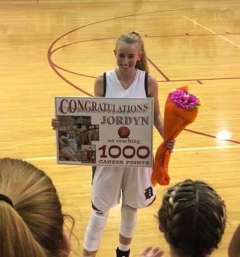 Female basketball player in a gym holding a bouquet of flowers and a congratulations yard sign.